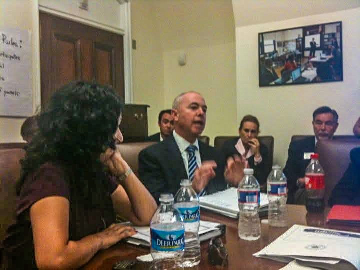 A crowded conference room, June 2012: USCIS Director Alejandro Mayorkas mid-sentence with both hands raised, Felicia Escobar listening in the near foreground, a flip chart of handwritten rules on the wall behind them, water bottles and papers across the table